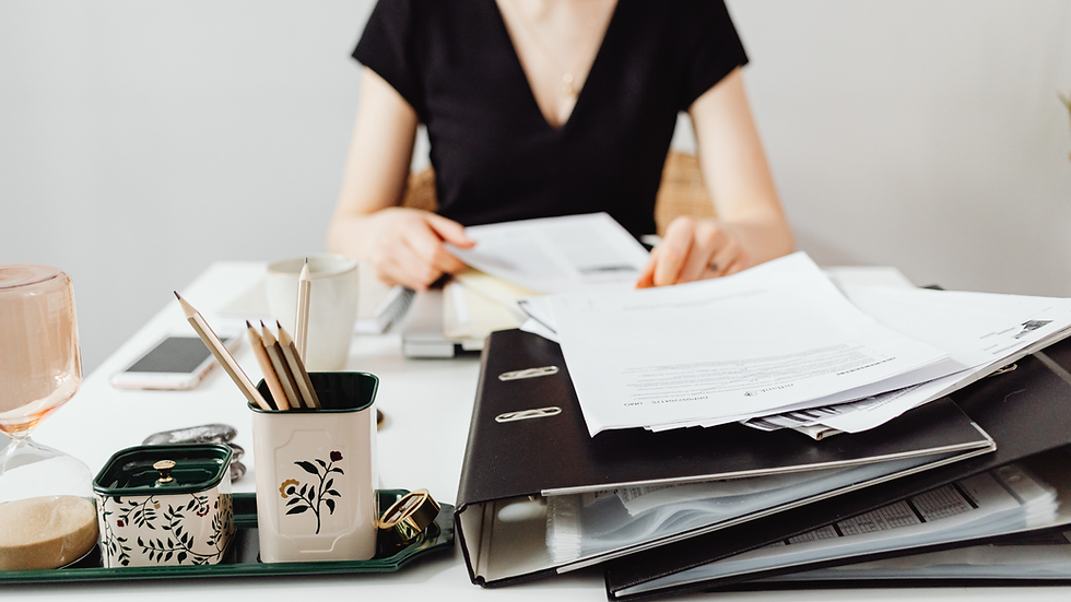 A person in a black shirt sits at a desk, reviewing documents. Papers, pencils in decorative holders, and a phone are on the white surface.