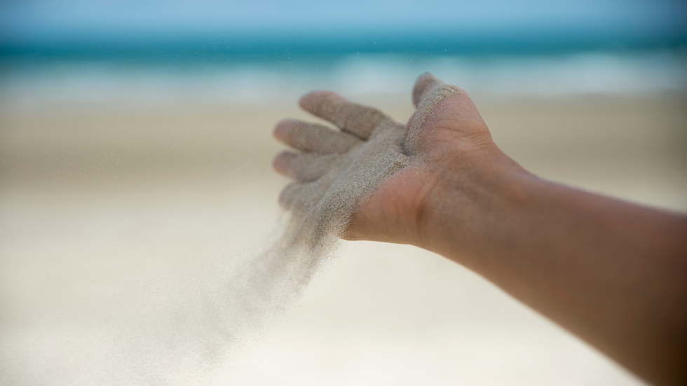 Hand releasing sand in the foreground with a blurred beach and ocean in the background; evokes a sense of calm and passage of time.