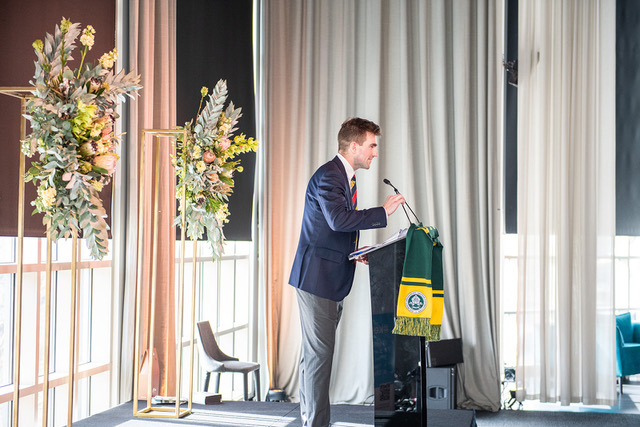 Man in suit speaks at a podium with a green and yellow scarf. Floral arrangements on stage. Bright, draped window background.