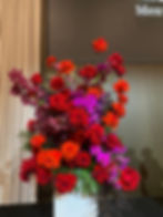 Vibrant stage floral arrangement with red roses, blood orange flowers, and purple orchids in a sleek vase. Curtain and shadowed wall backdrop.