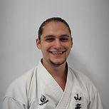 A smiling martial arts instructor wearing a white Tokaido gi with black Japanese characters and a logo on the chest, standing against a gray wall