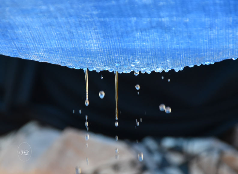 Sur le haut de la photo un tissu bleu bombé vers le bas. A l'arrière la nuit au et à l'arrière dans le bas de la photo des couleurs ocre et noir. Le tissu bleu est lourd de l'eau de la pluie et des gouttes tombent, d'autres s'échappent du tissu. Photo lumineuse et apaisante.