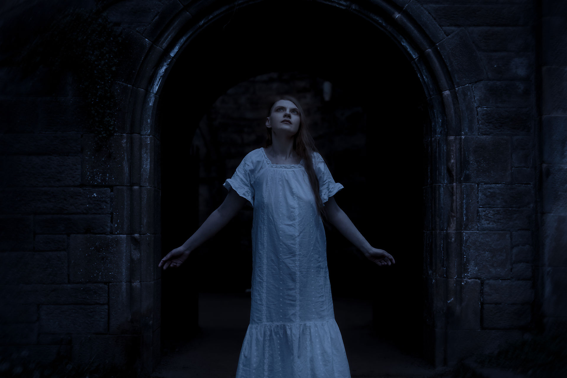Anastasia Dusk wearing a white nightdress stands with outstretched arms, gazing upwards in a dark, stone archway, evoking a gothic and ethereal presence. Inspired by Nosferatu.