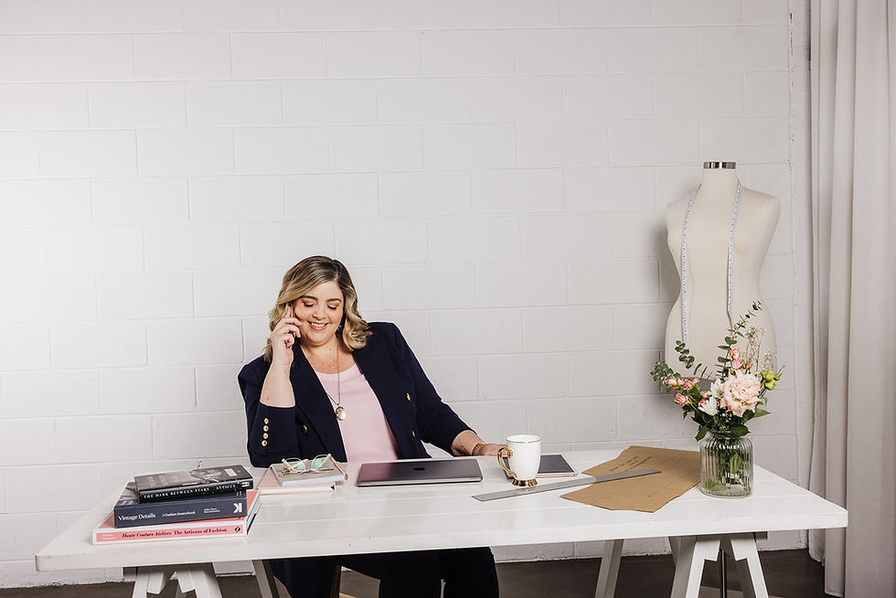 Smiling woman in navy blazer at a white desk with laptop, books and coffee mug, in a bright office with a white brick wall - Mel Rose Couture