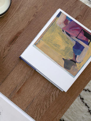 Art book on wooden table, abstract yellow and pink painting on cover, beside a sketchbook. Light background with a modern vibe.
