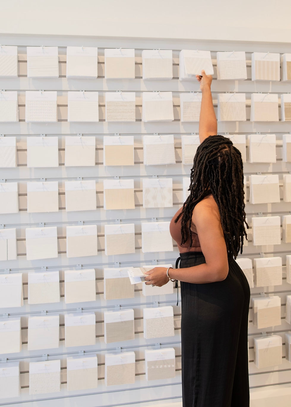 Interior designer selects a fabric sample from a display wall filled with swatches in a brightly lit showroom, holding multiple samples.
