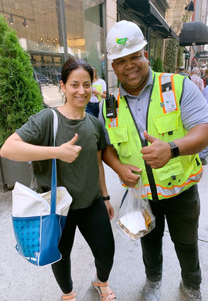 A woman and man on a city sidewalk. Woman carries a large bag on her shoulder and in other hand gives a lunch bag to a man wearing a construction helmet and yellow vest. Both are smiling, giving a 'thumbs up' sign.