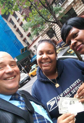 A man and two women on a city street, all smiling. Woman in center is wearing USPS sweatshirt, holding cash and pay it forward 9/11 smile card.