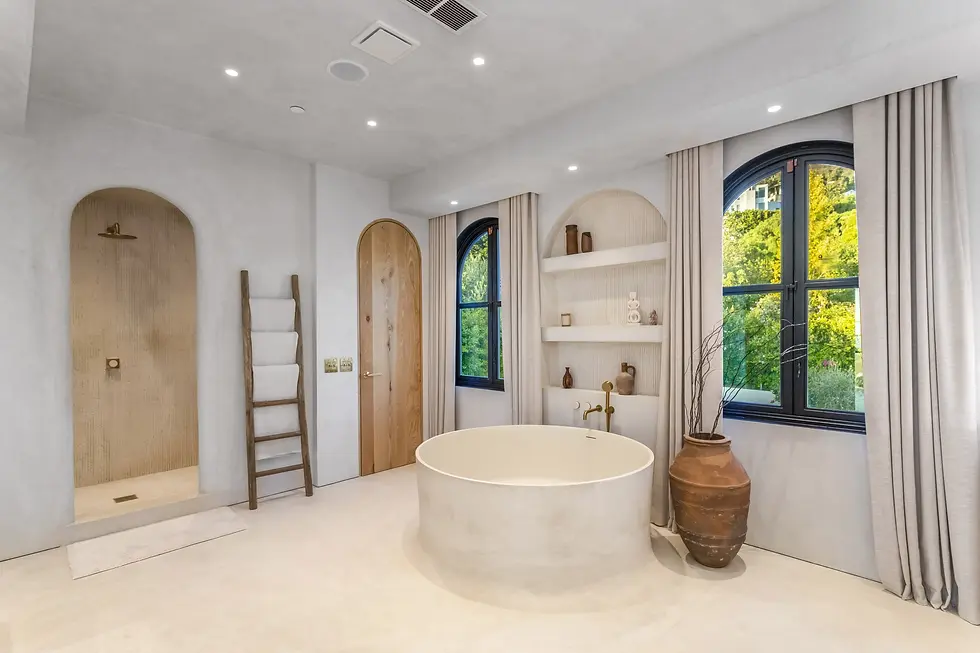 Modern spa-like bathroom with round soaking tub, arched shower, built-in shelves, black windows, and beige curtains overlooking green trees.