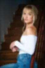 Debs Howard in white off-shoulder top and jeans stands by wooden stairs, looking at the camera with a calm expression.