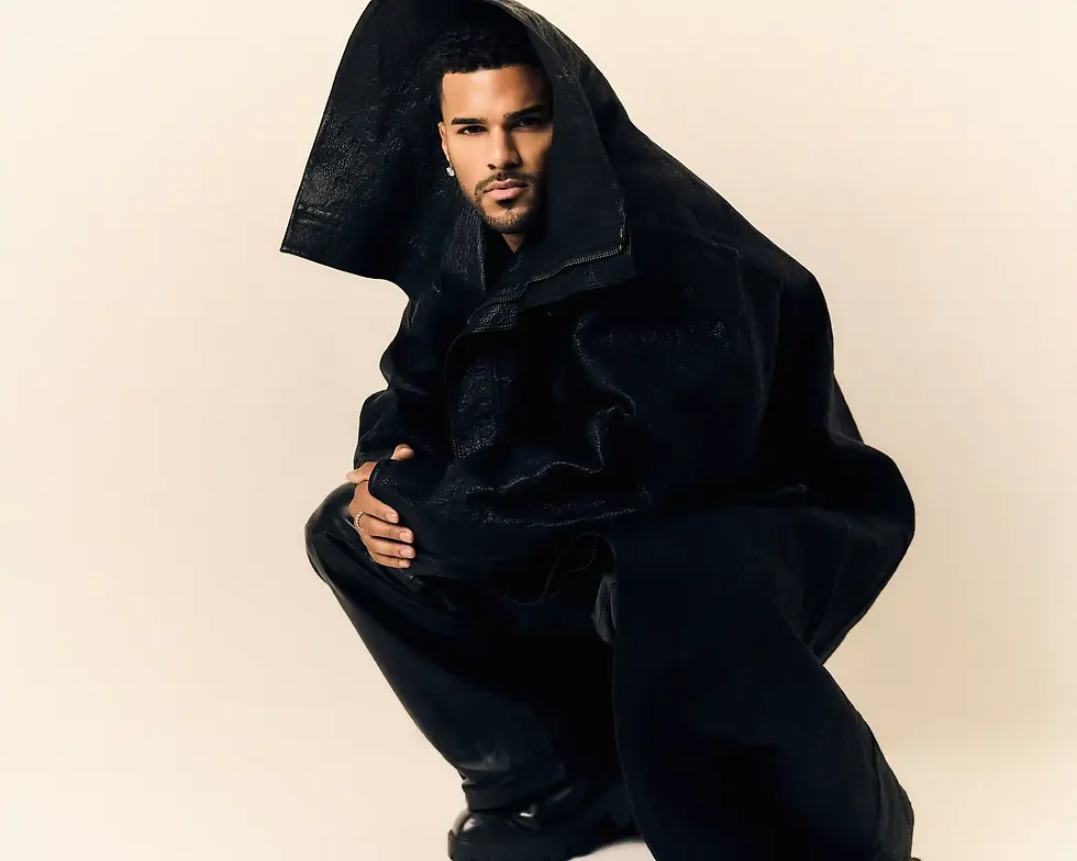 Jeremiah Brown in a black hooded outfit crouches against a plain beige background, posing like a fashion portrait.