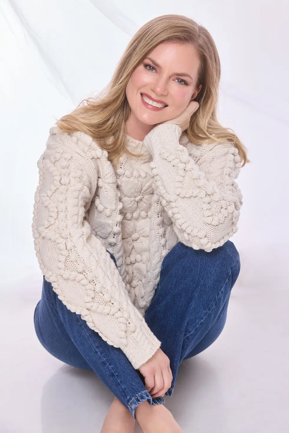 Cindy Busby in a cream textured sweater and blue jeans sits cross-legged against a white studio background.
