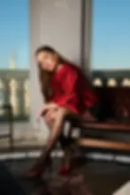 Actress Yağmur Yüksel in red jacket and polka-dot tights sits thoughtfully on a balcony with a cityscape view. Sky is clear and blue. Elegant mood.