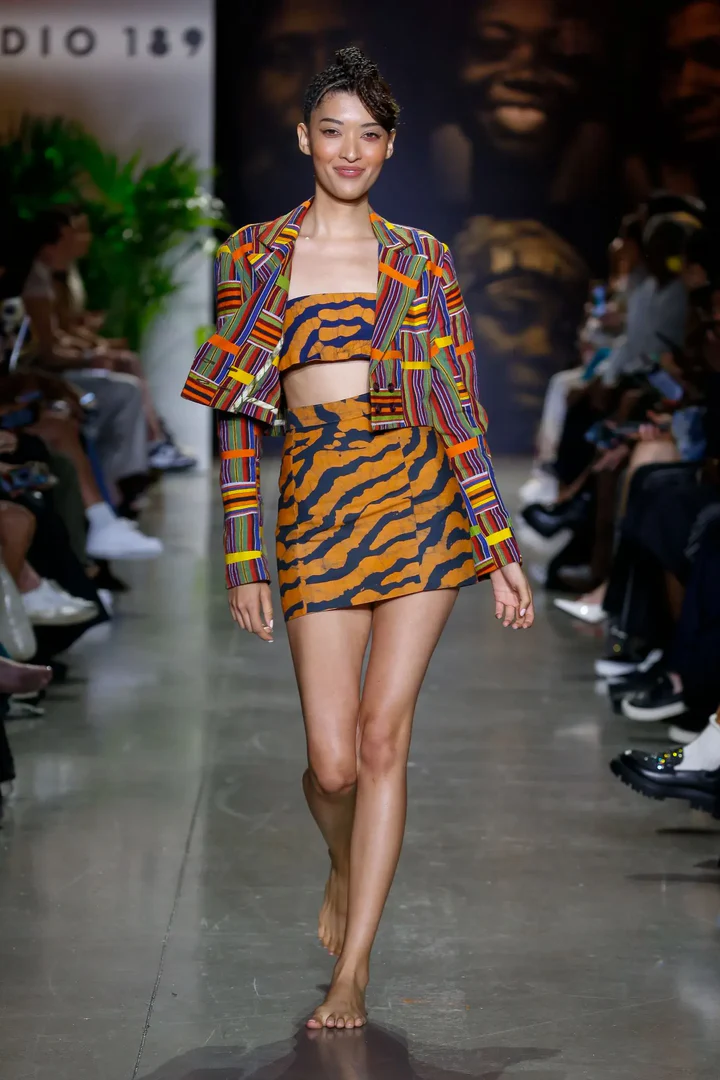 Model on runway displaying a vibrant striped blazer over a tiger print crop top and mini skirt, exuding bold style and confidence.