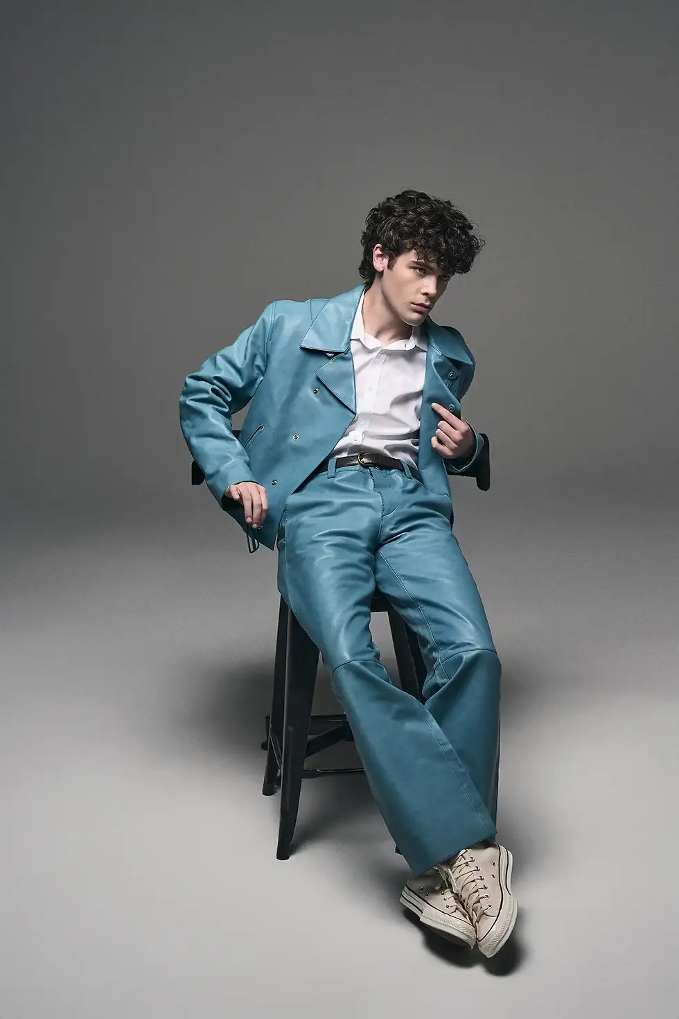 Duncan Joseph with curly dark hair in a bright blue jacket and white shirt, standing against a plain white background. in a blue suit and white shirt lounges on a black stool in a gray studio, looking serious.