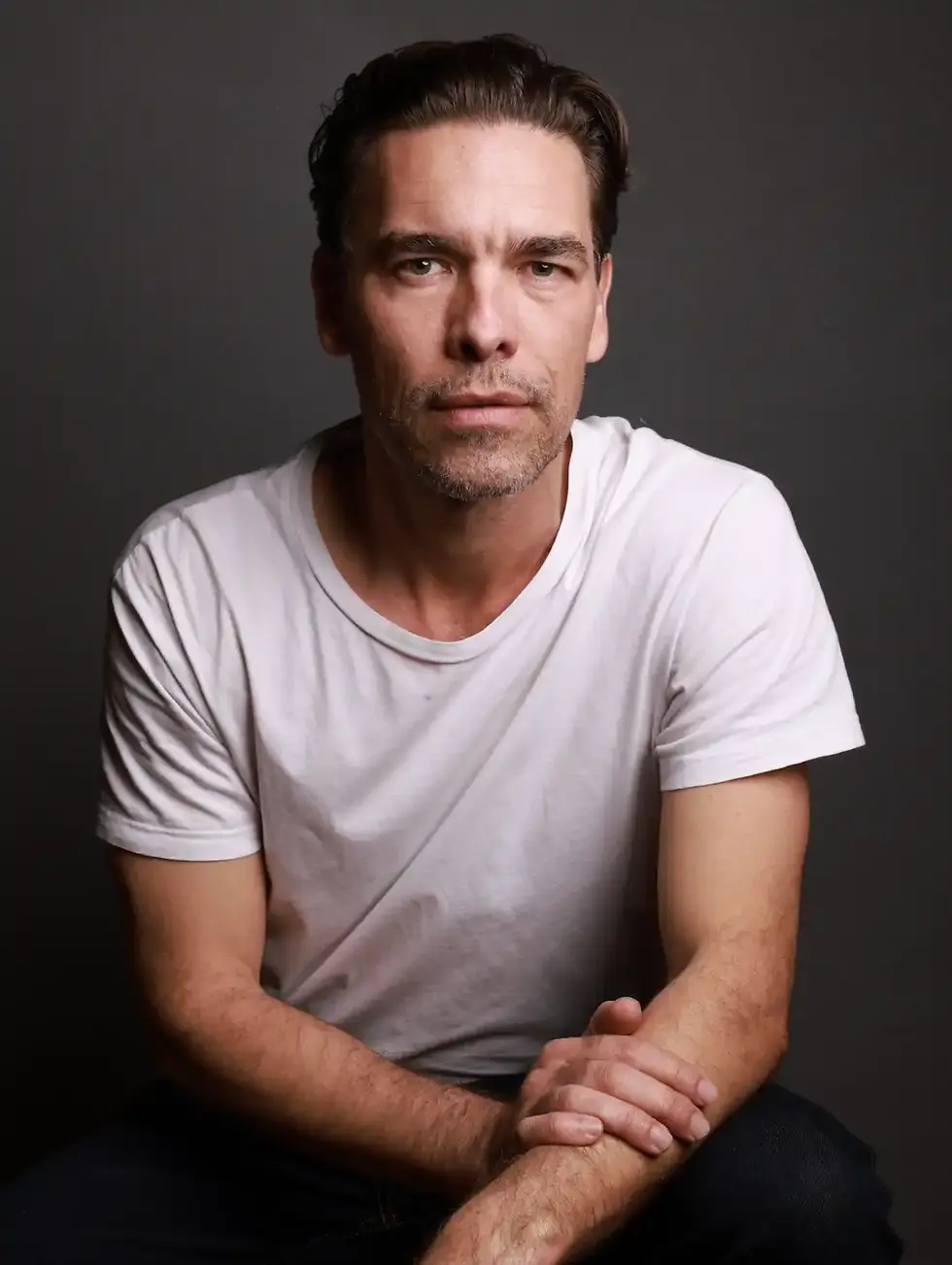 Thomas La Barbera Christensen in a white T-shirt, seated against a dark studio background, hands clasped.