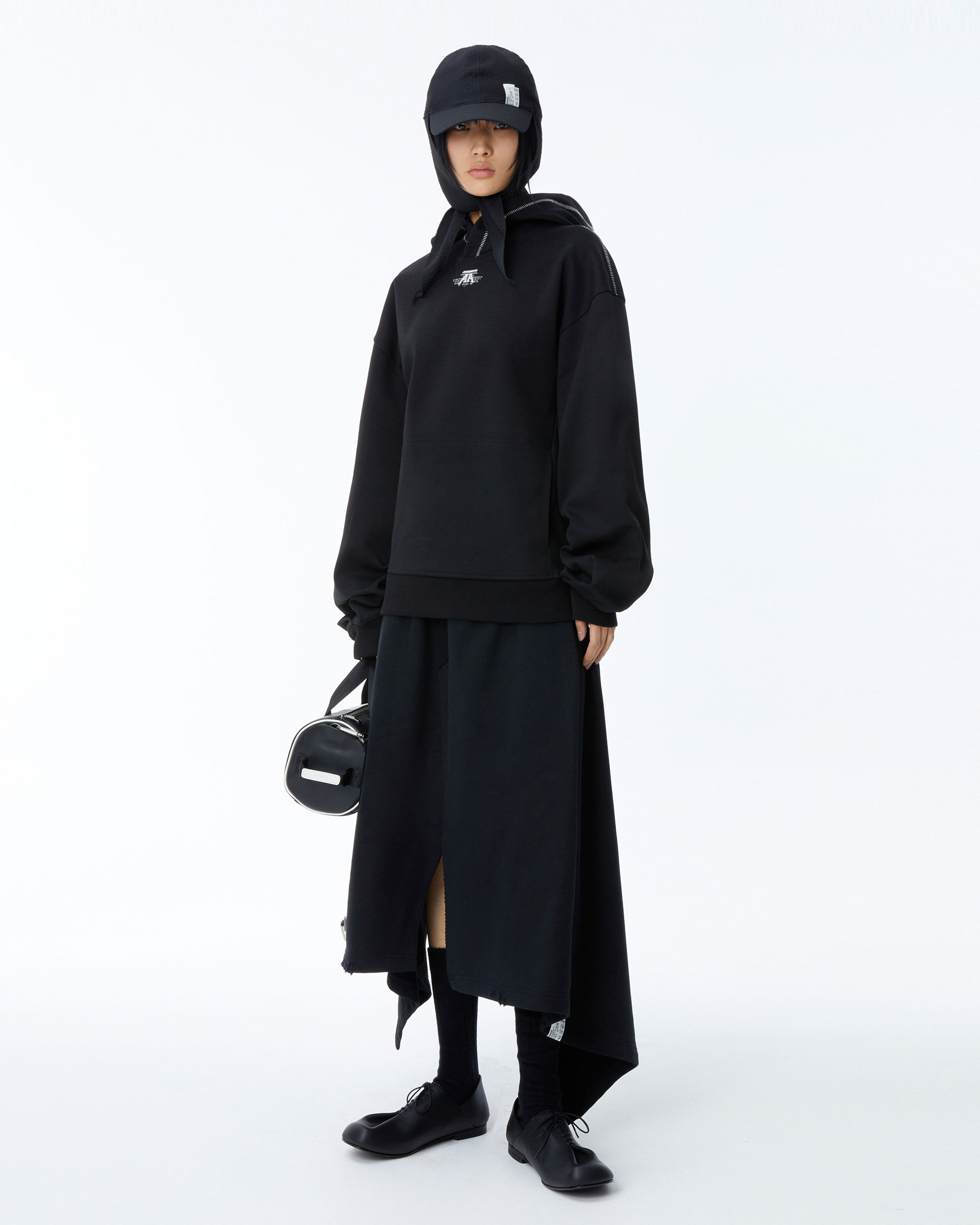 Model in a contemporary all-black streetwear ensemble featuring a hoodie, long skirt, and black shoes, with minimalist accessories.