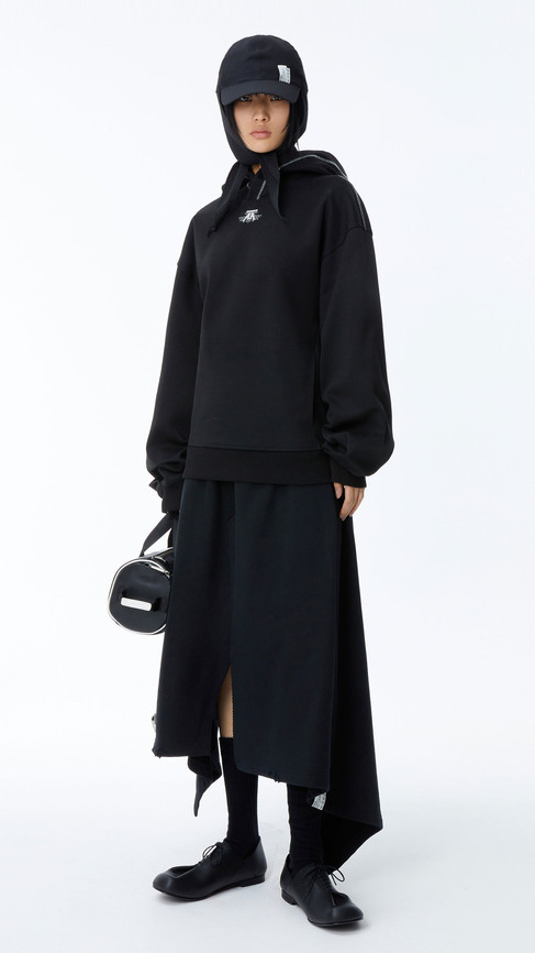Model in a contemporary all-black streetwear ensemble featuring a hoodie, long skirt, and black shoes, with minimalist accessories.