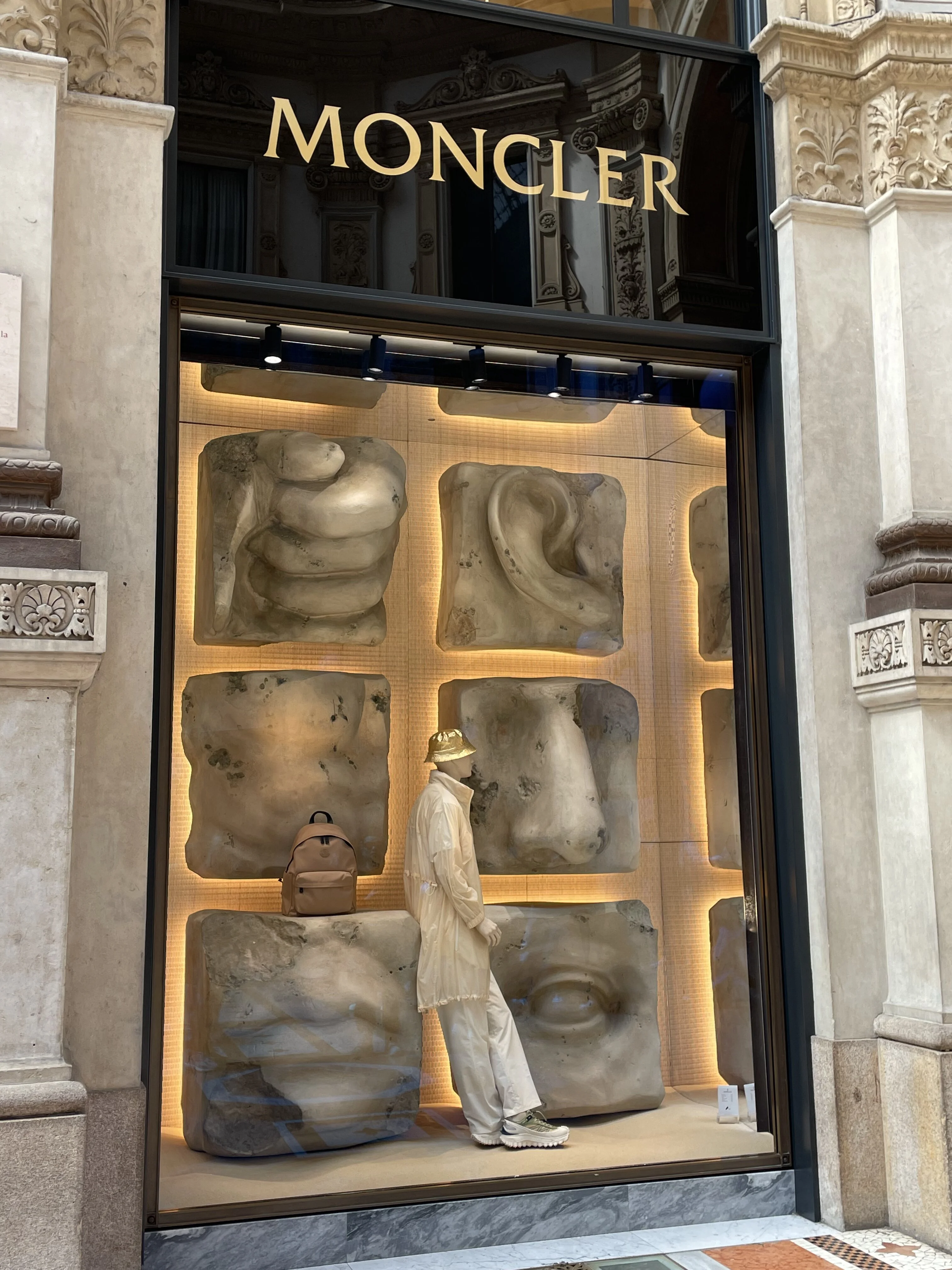 A Moncler store display with abstract human forms and designer bags in a window setup.