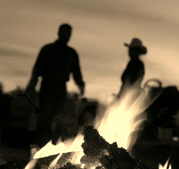 Studio or candid photography showing two silhouetted figures standing behind a bright, glowing campfire. The scene creates a warm, nostalgic, and peaceful atmosphere, reminiscent of an evening in the American West.