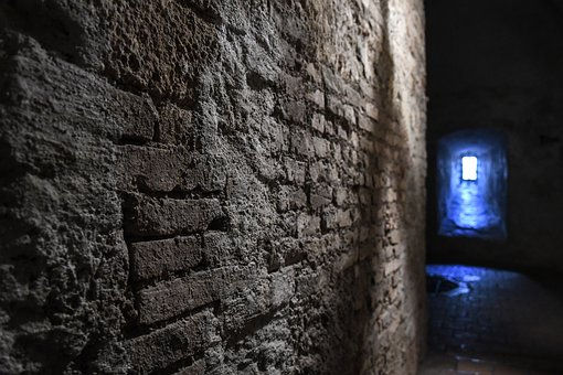 Picture of a gray brick wall leading down a hallway of a dungeon. There is a light at the end of the hallway