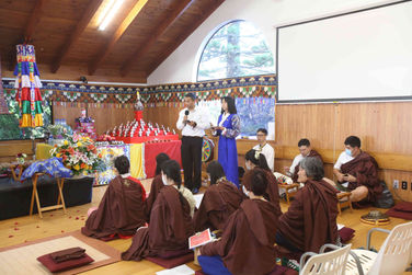 Two MCs stand before seated practitioners at the 2024 100,000 Ganachakra Offerings Ceremony