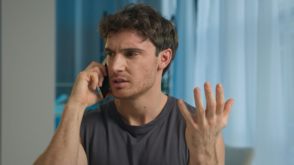 Man in gray shirt talking on phone, gesturing with one hand, looking frustrated. Blue curtains in the background create a calm setting.