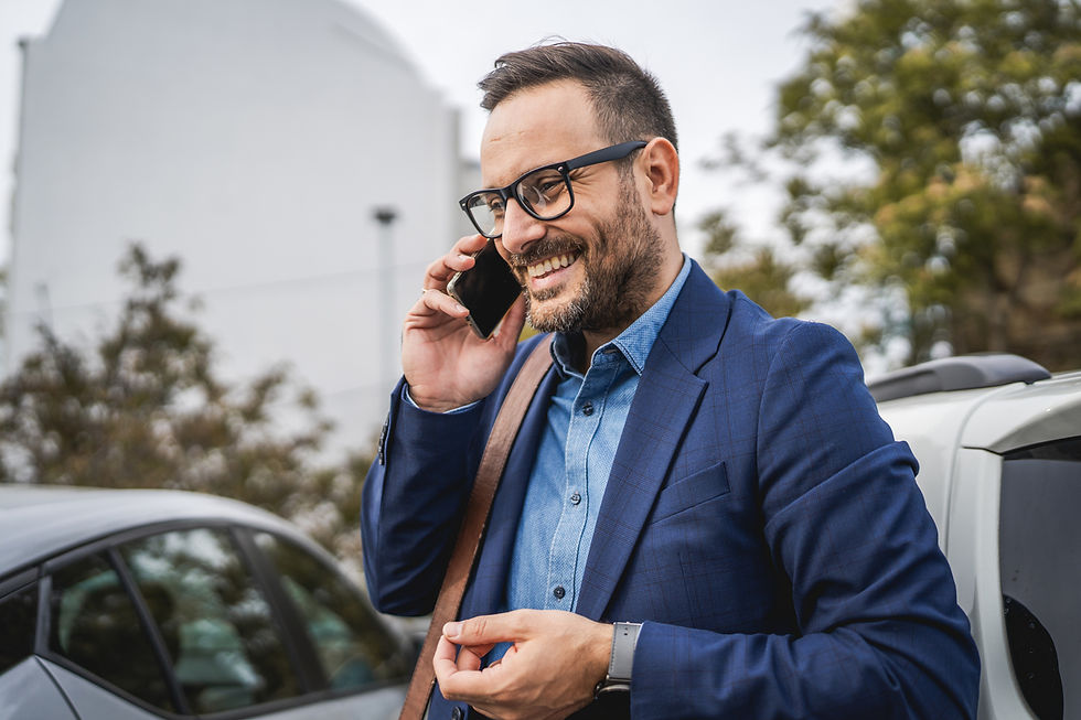 Smiling man in blue blazer talks on phone outdoors near a car, wearing glasses. Background features blurred trees and a white building.