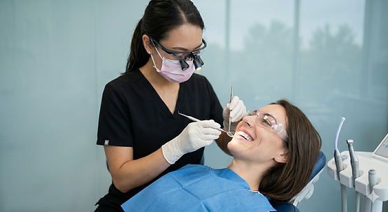 a dark-haired woman (dental patient) with a blue dental bib is leaning back while another dark-haired woman (dental hygienist) is performing a dental cleaning.