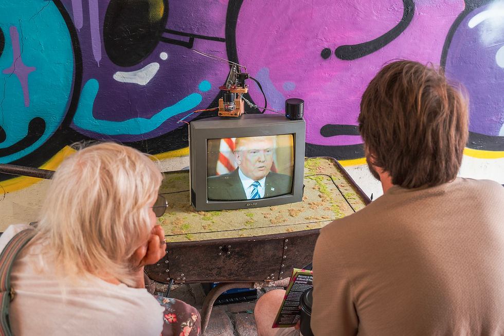 Two people watch a CRT TV showing a man speaking, set against a colorful graffiti wall. The scene is casual with a vintage vibe.