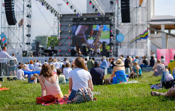 Outdoor music festival with people sitting on grass. Stage with large screen and lights in background. Relaxed and sunny atmosphere Space Coast Key Lime Pie Festival