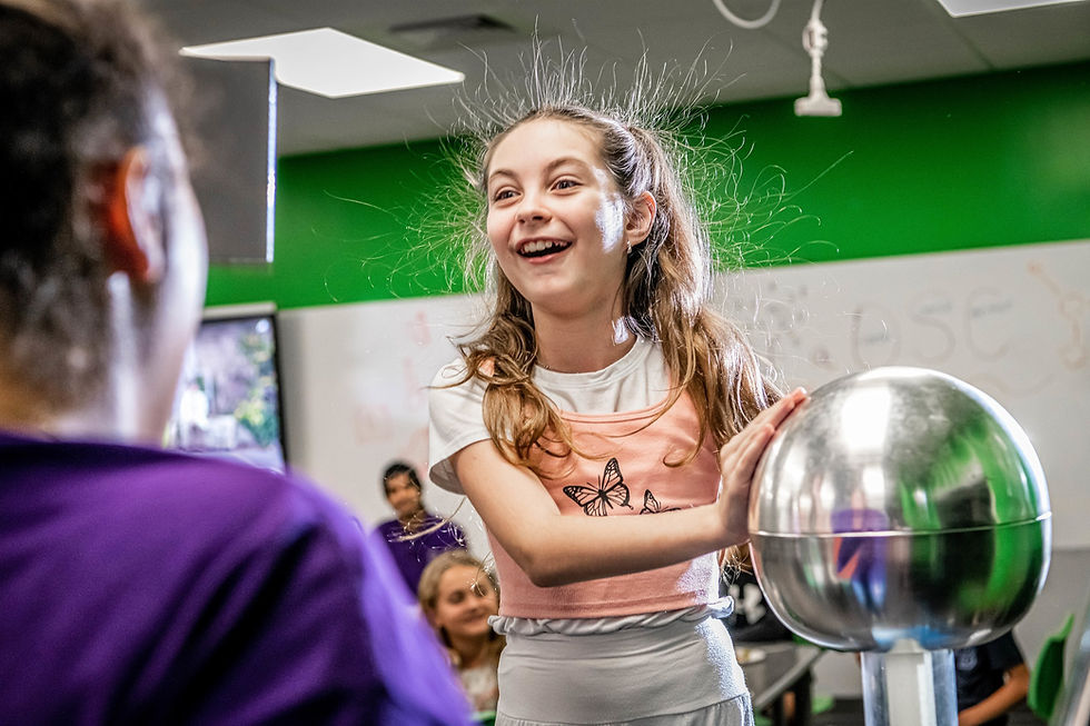 Girl with static-charged hair smiles, touching a metallic ball in a classroom. Green accent wall; others watch. Playful mood. Orlando Science Center Kids Night at the Museum