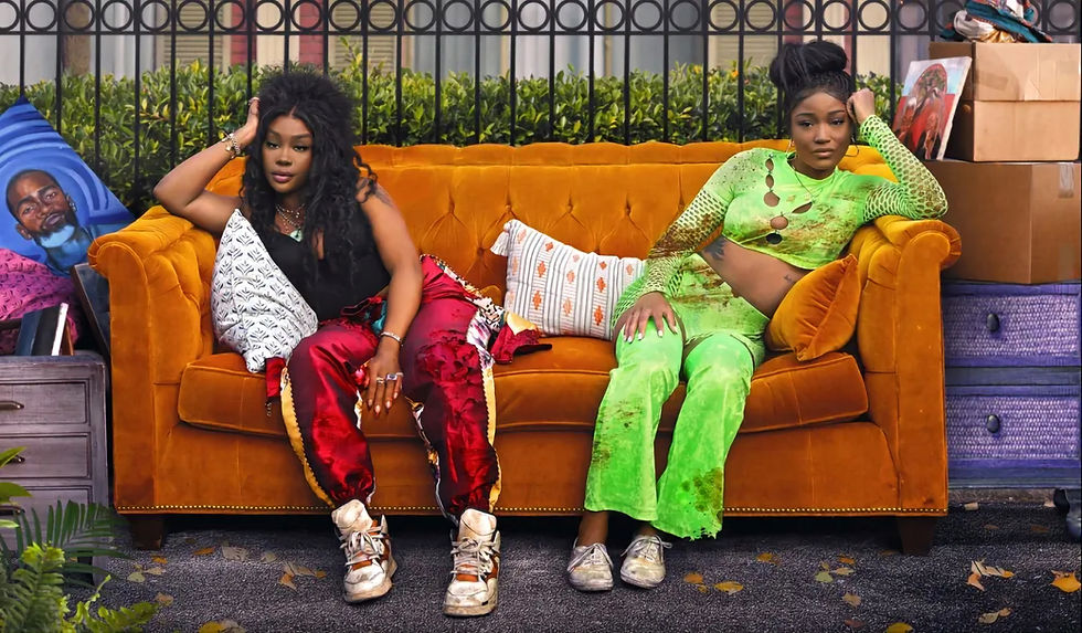 ONE OF THEM DAYS starring Keke Palmer and SZA. Two women sit on an orange couch outdoors. One wears red pants, the other a green outfit. Surroundings include plants and cardboard boxes.