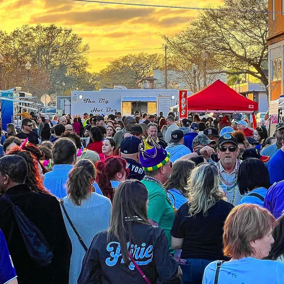 Crowd at a festive street event with food trucks and tents, colorful outfits, and a sunset backdrop. Lively atmosphere, text reads "The Big Hot Box." Sanford Mardi Gras Street Party