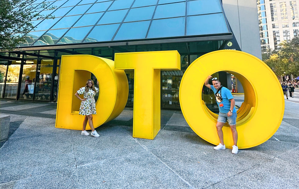 Gotta Go Emma and Shane posing with large yellow "DTO" letters in front of a glass building. Downtown Orlando DTO. Bright and cheerful atmosphere.