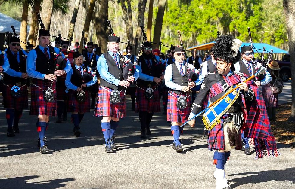 Bagpipers in red tartan kilts march on a sunny street, surrounded by trees. The leader wears a black feathered cap and blue sash. Central Florida Scottish Highland Games