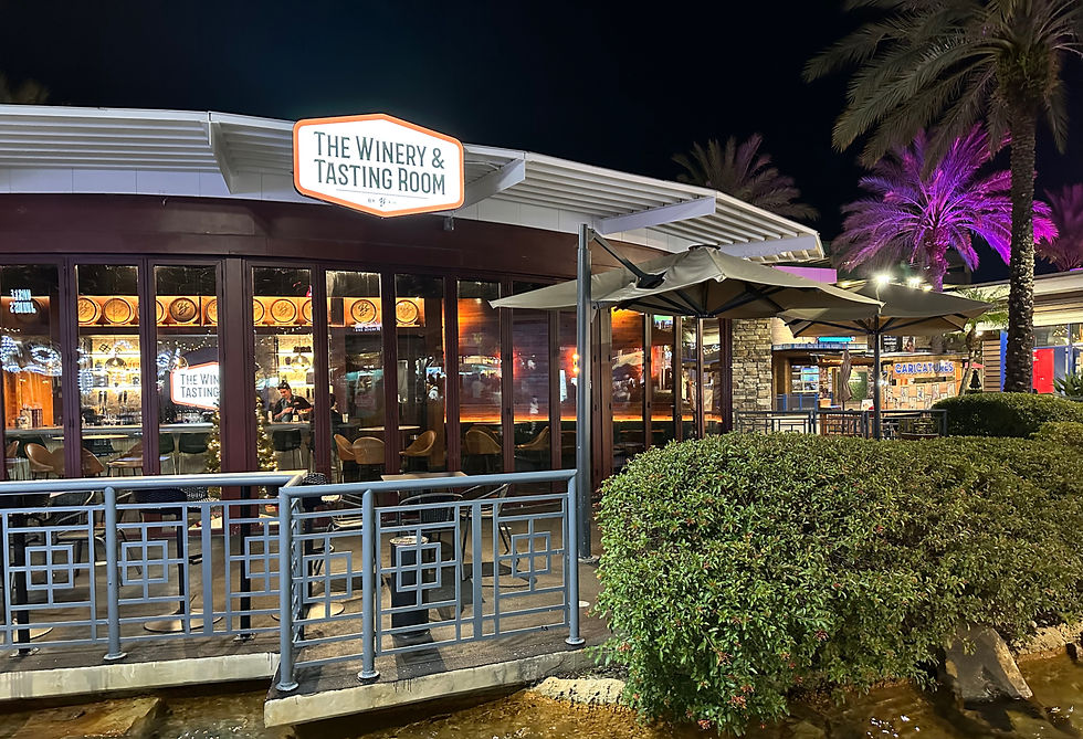Night view of a winery with warm lighting. Glass windows reveal the interior and a sign reads "The Winery & Tasting Room." Palm trees lit in purple. Located at ICON Park Orlando