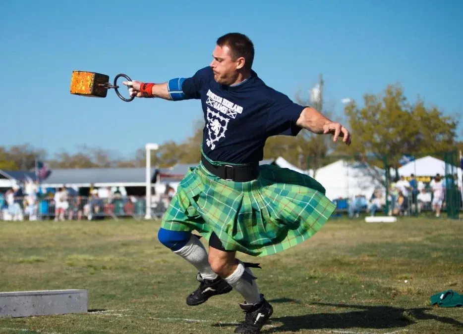 Man in green kilt throws weight in outdoor games. Wearing a navy shirt, dynamic pose, clear blue sky, lively fairgrounds in background. Mount Dora Scottish Highland Festival