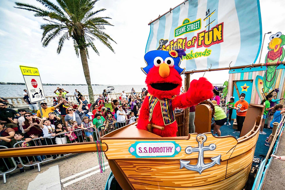 Elmo in a pirate costume waves from a float in a parade. Crowds cheer under a palm tree. The float features "S.S. Dorothy" and Sesame Street signage. Children’s Gasparilla in Tampa