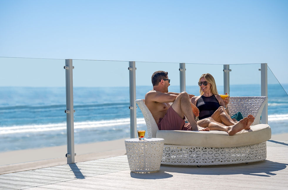 Couple relaxing on a patio daybed with drinks, overlooking a sunny Daytona Beach. They appear happy, surrounded by a clear blue sky and ocean.