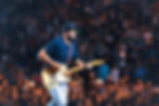 Musician in a blue shirt and cap plays electric guitar on stage. Energetic crowd in the blurred background, creating a lively atmosphere. Luke Bryan returns to Orlando's Kia Center on June 13 for the Country Song Came On Tour