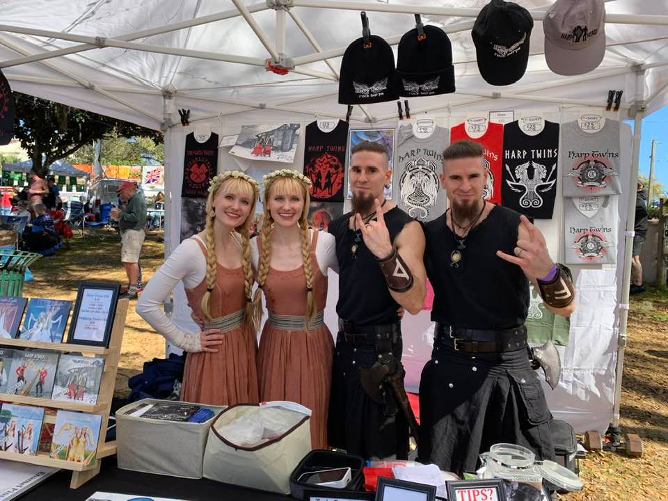 Festival booth with four people posing. Two women in long dresses, two men in dark attire. Merchandise includes CDs, shirts, and hats. Outdoor setting. Mount Dora Scottish Highland Festival