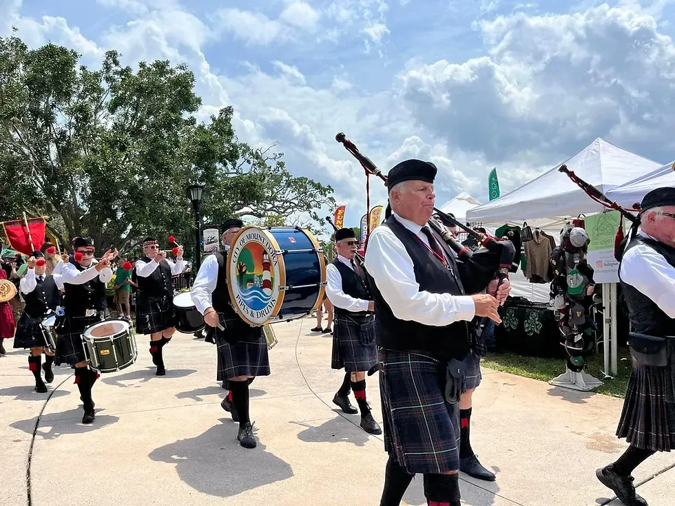 Bagpipers in kilts play in a parade under a cloudy sky, flanked by a drum with "City of Mount Dora" logo. Trees and tents in background. Mount Dora Scottish Highland Festival