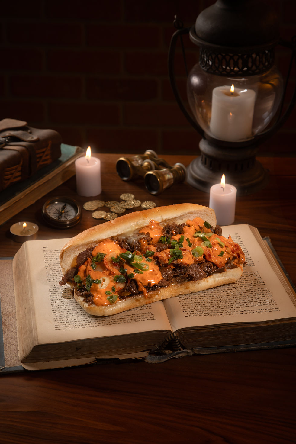 Sandwich with sauce and herbs on an open book, surrounded by candles, coins, a compass, and binoculars, creating a cozy atmosphere. Orlando Food Truck, Pacchioni's World Inspired Cheesesteaks