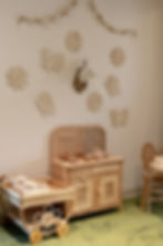 Children's play kitchen with wicker elements, rattan teapot and cups. Beige floral and butterfly wall decor against a soft green carpet. Cozy vibe. The Burrow Cafe and Play Mills 50 District