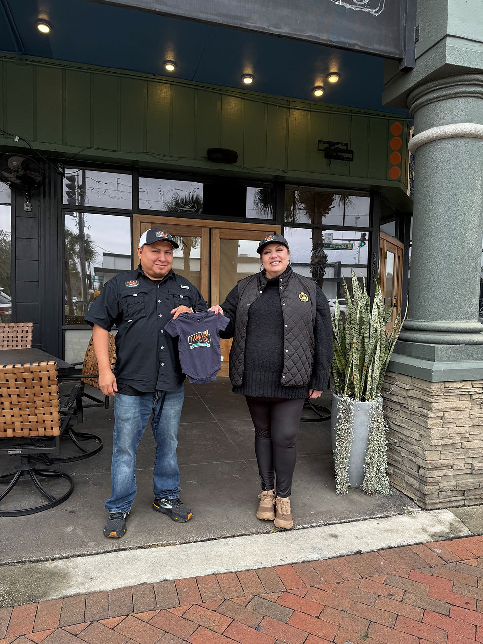 Two people smiling, one holding a navy shirt with "TAMACUN" text. They stand outside a green building with chairs and plants nearby. Tamale Co. Modern Mexican Kitchen & Bar opening new restaurant in College Park