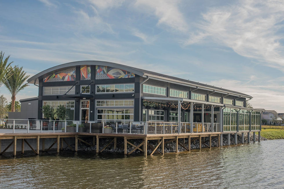 Modern waterfront building with colorful mural, glass windows, and outdoor seating. Palm trees and blue sky create a serene atmosphere. Canvas Restaurant Lake Nona