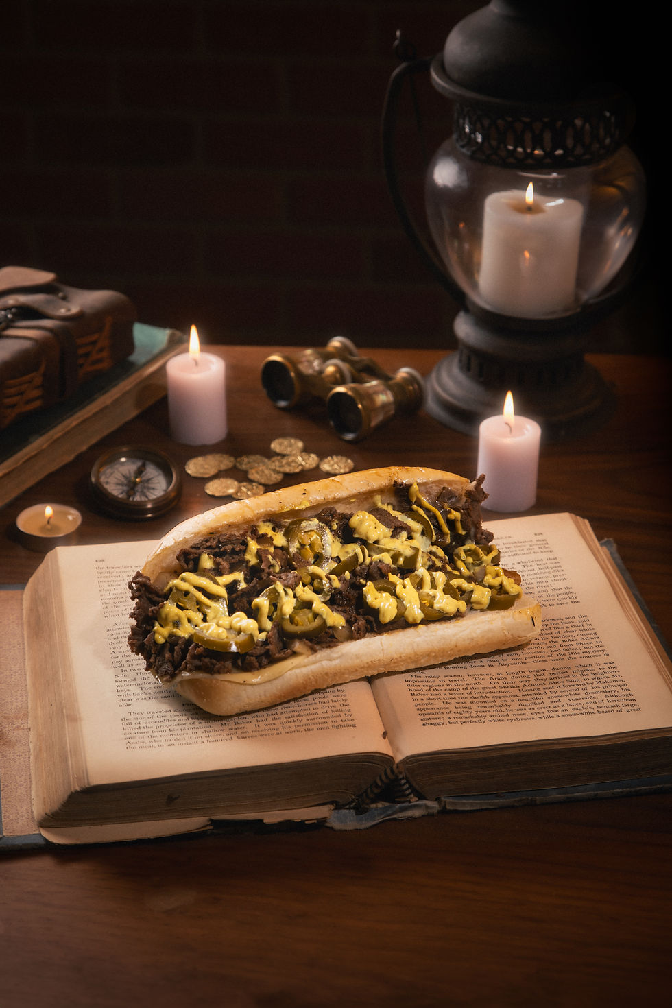 Cheesesteak with mustard on open book, surrounded by lit candles, a compass, and binoculars on wooden table, creating a vintage vibe. Orlando Food Truck, Pacchioni's World Inspired Cheesesteaks