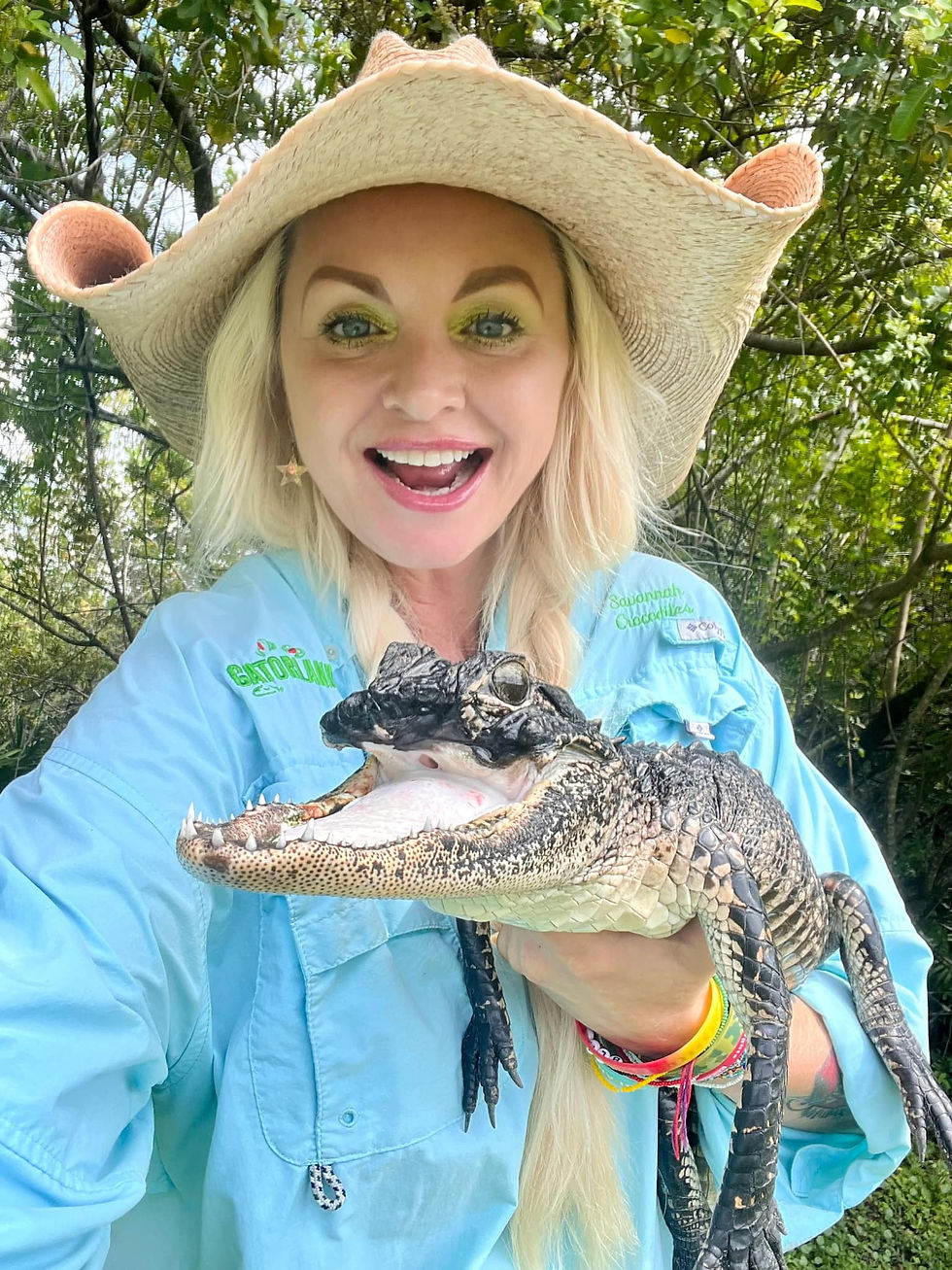 A smiling Savanna in a cowboy hat holds Jawline, an alligator. She's wearing a blue shirt with "Gatorland" text. Green foliage background.