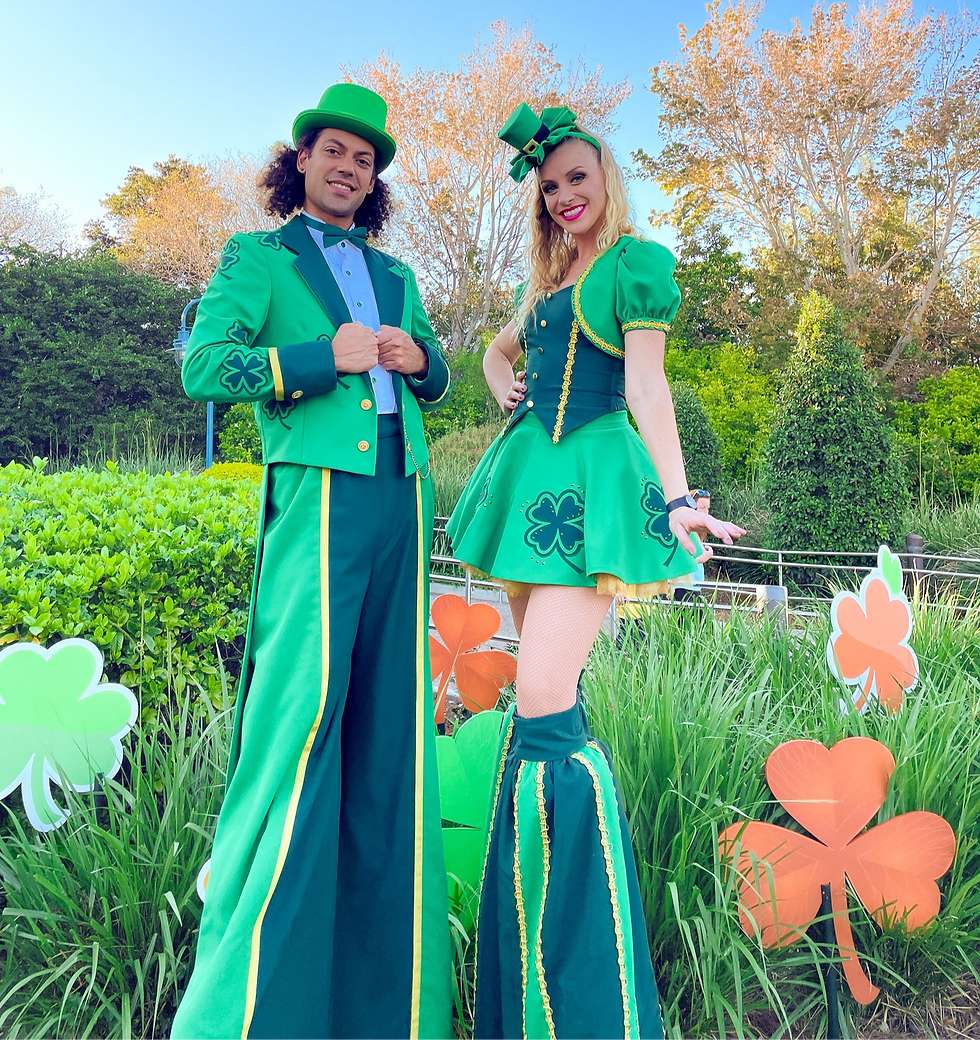 Two people on stilts dressed in green outfits with shamrocks smile in a garden setting. Bright foliage and orange shamrocks in background. St. Patrick's Day at SeaWorld Orlando